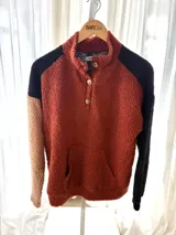 Buzo de corderito con diseño colorblock en tonos ladrillo, negro y beige, con cuello alto, cierre frontal parcial con botones y bolsillo tipo canguro.