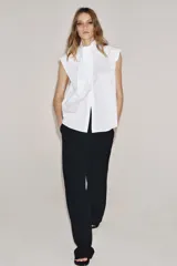 Camisa sin mangas confeccionada en algodón, con cuello redondo y detalle de lazada frontal. Presenta cierre frontal con botones ocultos por solapa y bajo redondeado.