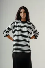 Camiseta de manga larga, corte holgado, con cuello redondo y estampado de rayas horizontales anchas en blanco y negro. Presenta un pequeño bordado rojo en el pecho.