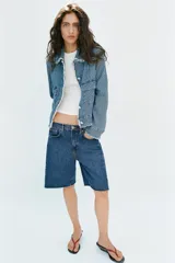 Bermuda de denim azul de tiro medio y corte relaxed, con diseño de cinco bolsillos y bajo desflecado. Cuenta con cierre frontal de cremallera y botón metálico.