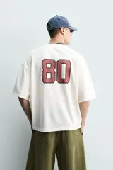 Remera de algodón color crudo, con textura de tejido abierto, cuello redondo y mangas cortas. Presenta estampas en contraste en el frente con los textos 'Field Club', 'West Coast', gráficos deportivos y un escudo circular.