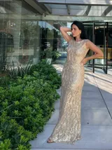 Vestido largo sin mangas, color dorado, con base beige y diseño de encaje o brocado con brillo. Corte ajustado al cuerpo que cae recto hasta el suelo.