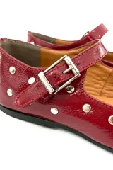 Zapato tipo Mary Jane color cereza charolado, con tachas plateadas, correa con hebilla y suela plana.