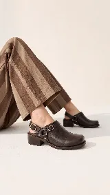 Zueco de cuero marrón oscuro, con diseño cerrado en la parte delantera, tira ajustable en el talón con hebilla metálica y taco bajo cuadrado.