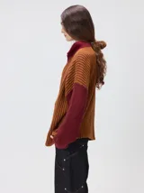 Pullover de tejido acanalado grueso con cuello alto y cierre frontal parcial. Presenta un diseño de bloques de color, con cuerpo en tono beige y mangas en contraste con rayas verticales en tonos rojizos. Incluye un detalle bordado en el pecho.