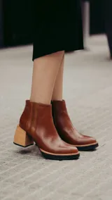 Bota corta de cuero color marrón con acabado liso, que presenta un detalle de textura acanalada en el lateral y cierre interno. Cuenta con un taco ancho y estable de color negro.
