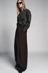 Pantalón ancho de tiro medio color marrón oscuro, confeccionado en tejido con lana. Presenta pinzas delanteras, cinturón ajustable con hebilla metálica y bolsillos cargo de plastrón en las piernas.