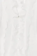 Camisa de manga larga en color blanco, con cuello redondo, escote en pico y cierre frontal con botones. Presenta detalles de volantes en el mismo tejido a lo largo de la abertura frontal.