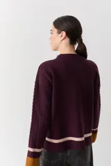 Buzo de punto color crudo con mangas de textura trenzada, detalles en contraste en los puños y franjas horizontales en el ruedo. Presenta cuello redondo y un pequeño bordado en el pecho.