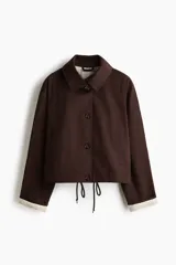 Chaqueta corta de sarga marrón con cuello camisero, manga raglán y cierre frontal de botones. Cuenta con bolsillos laterales y cordón ajustable en el bajo, con detalles de forro a rayas en los puños y el interior.