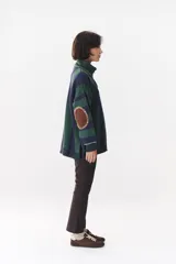 Buzo de punto con diseño de rayas verticales en tonos verde y azul marino. Presenta cuello alto con cierre frontal, hombros caídos y coderas en contraste de color marrón con costuras decorativas visibles.