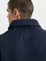 Cardigan de punto color marrón con cuello esmoquin, cierre frontal con botones y presilla superior. Presenta mangas largas con coderas en contraste y bolsillos frontales de ribete.