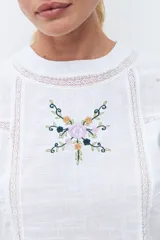 Blusa blanca de manga larga con cuello redondo, confeccionada en tela liviana con detalles de encaje vertical y bordado floral en el pecho.
