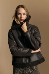 Campera tipo gamulán de cuero sintético negro, con interior y detalles de corderito sintético al tono en cuello y ruedo. Presenta cierre frontal metálico y diseño de silueta relajada.