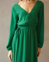 Vestido largo de satén color verde esmeralda, con escote cruzado, mangas largas amplias con puños elásticos y cintura marcada con lazo ajustable.