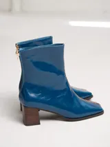 Botinetas de cuero azul con acabado charol, diseño de punta cuadrada, taco cuadrado de madera y cierre en la parte trasera. Presentan una costura bordada en color contrastante a lo largo del empeine.