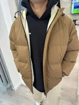 Campera puffer de color marrón claro con diseño acolchado, cierre frontal completo y capucha ajustable con cordones. Cuenta con forro interior en color crema y bolsillos laterales.