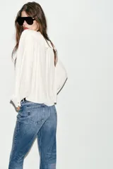 Blusa de georgette en color blanco, con escote en pico, detalle de volantes y lazada frontal. Presenta mangas largas abullonadas con puños ajustados y un bajo asimétrico con diseño tipo globo.