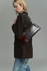 Bolso de hombro de formato alargado, color marrón oscuro con acabado ligeramente desgastado. El asa de hombro es delgada y ajustable mediante hebillas metálicas. Presenta detalles de tachas o remaches metálicos en los puntos de unión del asa.