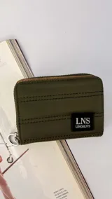 Billetera corta de nylon color verde militar, con diseño capitoneado y cierre perimetral. Incluye una etiqueta frontal con el logo de la marca LNS Lincoln's y una argolla metálica lateral.