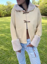 Campera de gamuza color beige con interior y detalles de corderito sintético en cuello y puños. Presenta cierre frontal con botones tipo toggle de madera y cuero, y bolsillos frontales.