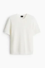 Remera de punto color blanco, con textura de pequeños patrones en relieve, cuello redondo y mangas cortas.