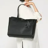 Cartera tipo tote bag Guess modelo Lucci, de color negro con acabado texturizado símil cuero. Cuenta con doble asa superior, herrajes dorados, placa metálica triangular con el logo de la marca y un charm colgante en forma de flor.