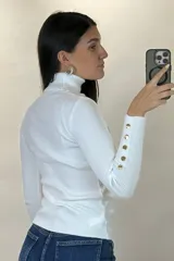 Polera de hilo blanca de cuello alto y manga larga, con detalle de botones dorados en los puños. Tejido de punto acanalado ajustado al cuerpo.