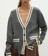 Cardigan de punto gris con escote en V, cierre frontal con botones y detalles de vivos en color crema en el escote, puños, bolsillos y ruedo. Presenta un diseño de corte holgado con mangas largas y bolsillos funcionales.