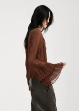 Blusa de color marrón chocolate, confeccionada en tejido plisado con textura. Presenta un diseño de cuello redondo con detalle de lazo, mangas largas amplias con puños acampanados y un corte holgado con volados sutiles en la parte frontal.