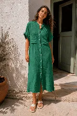 Vestido camisero midi de lino verde oscuro, con cuello mao, mangas cortas con vuelta y cierre frontal de botones de nácar. Presenta bolsillos appliqués en el pecho y un corte recto y holgado.
