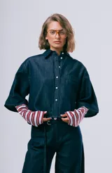 Camisa de denim azul oscuro, de corte corto y oversize, con mangas amplias de largo intermedio, cuello camisero, cierre frontal con botones y un bolsillo aplicado en el pecho.