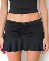 Minifalda negra con diseño fruncido en la parte central delantera, detalle de lazo pequeño en la cintura y borde con terminación de encaje. Presenta un ruedo con volado suave.