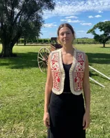 Chaleco color crudo, sin mangas, con bordados artesanales en tonos rojo óxido y detalles geométricos y botánicos. Presenta ribetes plisados a lo largo de la abertura frontal.