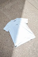 Remera blanca de manga corta con cuello redondo, pequeña estampa frontal y etiqueta decorativa en el borde inferior.