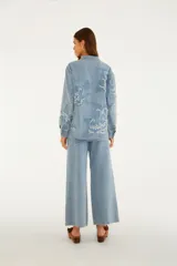 Pantalón jean de tiro medio y corte recto, color azul claro, con grandes bordados florales en tono sobre tono y detalles deshilachados. Cuenta con bolsillos funcionales y cierre frontal con botón y cremallera.