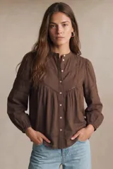 Blusa de algodón color marrón con cuello mao, cierre frontal con botones y mangas largas con puños elásticos. Presenta detalles de encaje y bordados en el canesú y en las mangas, con un diseño holgado y fluido.