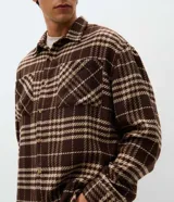 Camisa tipo overshirt de corte oversized, confeccionada en tejido de poliéster con textura. Presenta un estampado de cuadros en tonos marrones y beige, cuello clásico, cierre frontal con botones y bolsillo aplicado en el pecho.