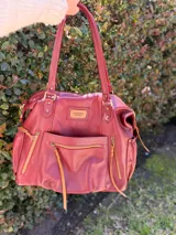 Bolso maternal de cuero marrón con doble asa de hombro, correa ajustable, múltiples bolsillos frontales y laterales con cierre, y detalles de tiras trenzadas.