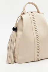 Mochila de símil cuero texturizado color beige, con asa superior, tiras regulables para la espalda y cierre principal metálico. Presenta un detalle trenzado vertical en el frente y un accesorio con flecos y cuenta negra en el lateral.