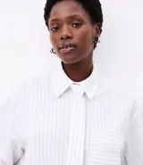 Camisa de mujer de corte maxi oversized, confeccionada en tricolina de algodón certificado. Presenta rayas verticales de hilo teñido, cuello con corbata (gravata), botones frontales, bolsillo en el pecho y puños largos.