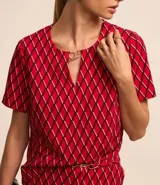 Blusa de crepé color rojo con estampado geométrico de rombos en negro y blanco. Tiene cuello redondo con abertura en forma de gota y detalle metálico, mangas cortas y corte recto.