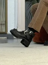 Botinetas de cuero negro con suela track, punta cuadrada y doble hebilla metálica decorativa en el lateral. Diseño de caña corta con cierre lateral.