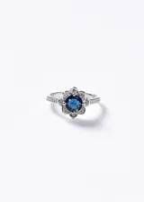 Anillo de plata 925 con diseño de flor central que presenta una piedra azul redonda rodeada de pequeñas zirconias blancas. El aro del anillo también cuenta con detalles de zirconias incrustadas.