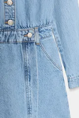 Vestido corto de denim azul claro, estilo camisa, con cuello solapa, escote pico, manga larga y cierre frontal de botones metálicos. Presenta bolsillos delanteros con tapa y bolsillos de plastrón en la espalda. La cintura está ajustada con elástico fruncido.