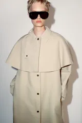 Trench de cuello con solapa y manga larga, con detalle de capa desmontable del mismo tejido en la parte superior. Cuenta con cierre frontal de botones y bolsillos en el delantero.