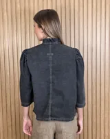 Camisa de denim color negro con efecto gastado, cuello alto con volados sutiles, mangas tres cuartos abullonadas en los hombros y cierre frontal con botones metálicos y costuras pespunteadas en contraste.