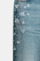Jeans tiro medio de corte relaxed leg y largo completo, confeccionados en denim azul claro. Presentan detalles de aplicaciones de pedrería y joyas a contraste en los laterales superiores y bolsillos delanteros, estilo clásico de cinco bolsillos y cierre frontal con cremallera y botón metálico.