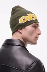 Gorro tipo beanie de punto color verde oliva con el logo de la marca Obey bordado en letras amarillas con contorno blanco en la parte frontal.