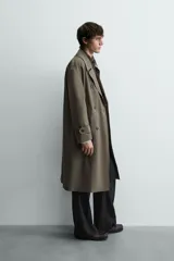 Trench color verde militar, de corte oversized, con cuello con solapas de muesca y manga larga con puño y trabilla. Presenta bolsillos de plastrón con solapa en la cadera y un bolsillo interior. Tiene una abertura en el centro de la espalda y cierre frontal de botonadura cruzada.